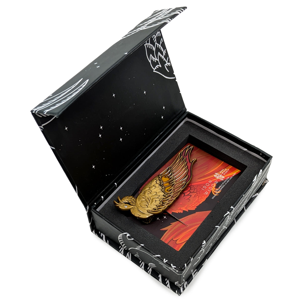 The Phoenix Hair Clip with its backing card nests perfectly inside its open, black box packaging. The white lineart continues onto the underside of the box as well as along the outer edges.