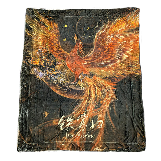 A flat lay product photo of the Phoenix Usurping Dragon Blanket. The blanket is illustrated with a screeching and fiery feathered phoenix rising in flight. The colors of the blanket are fiery muted orange and yellow of the phoenix on a warm grey background. At the bottom, Iron Widow with its Chinese characters are printed in gold. 