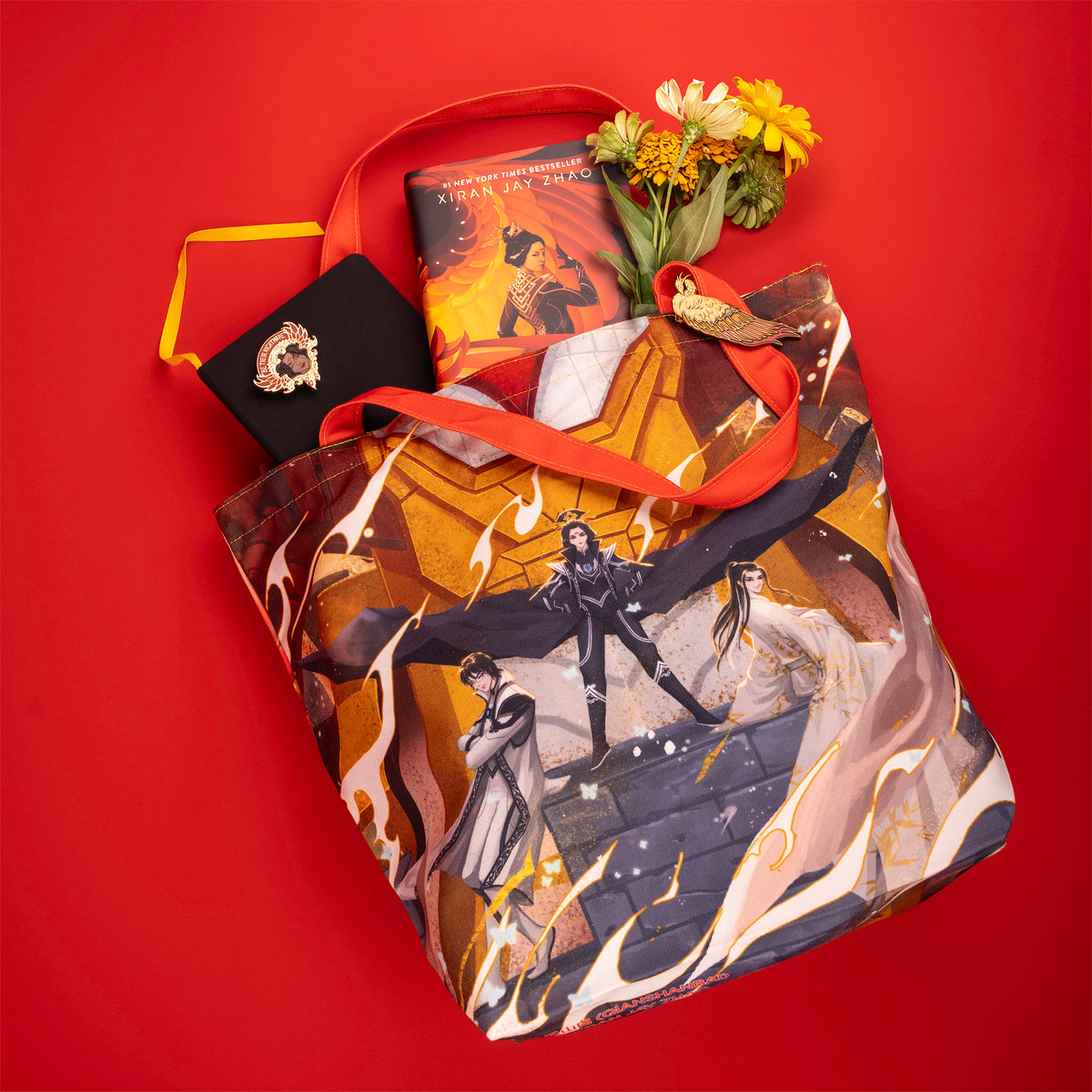 Iron Triangle Tote, which has an illustration of the Vermillion Bird’s face gritting its mecha teeth, lays flat on the red background. Different items are spilling out of it, including a copy of Iron Widow, flowers, and sunglasses. The Vermillion Bird Hair Clip is clipped to the tote’s shoulder strap.