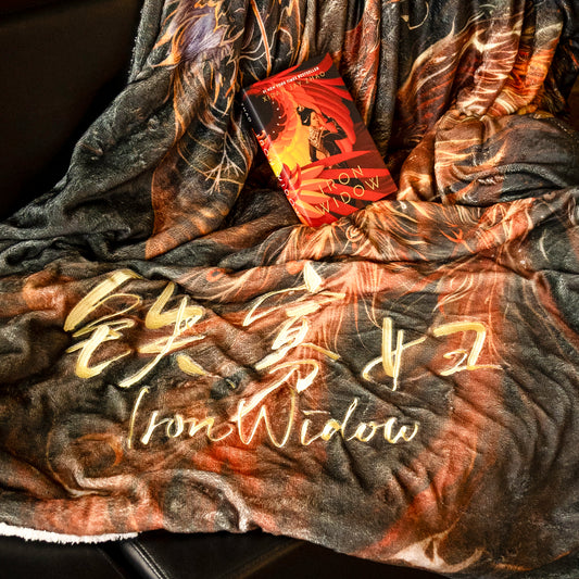 The Phoenix Usurping Dragon Blanket is thrown over the front of a sofa. The print of Iron Widow and Chinese characters in gold is showcased. The colors of the blanket are fiery muted orange on a warm grey background. The full illustration of a phoenix rising is obscured by the blanket wrinkles.