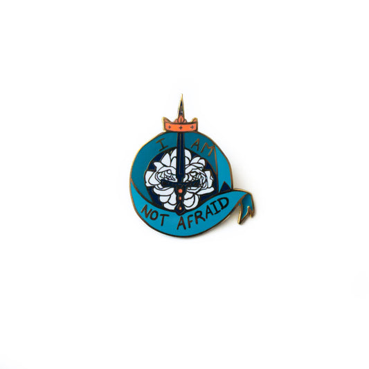  A product photo of an enamel pin depicting a flowing teal ribbon in a circle around a sword on a bed of unfurled white roses. A coral crown is placed below the tip of the sword. The ribbon reads “I Am Not Afraid.”