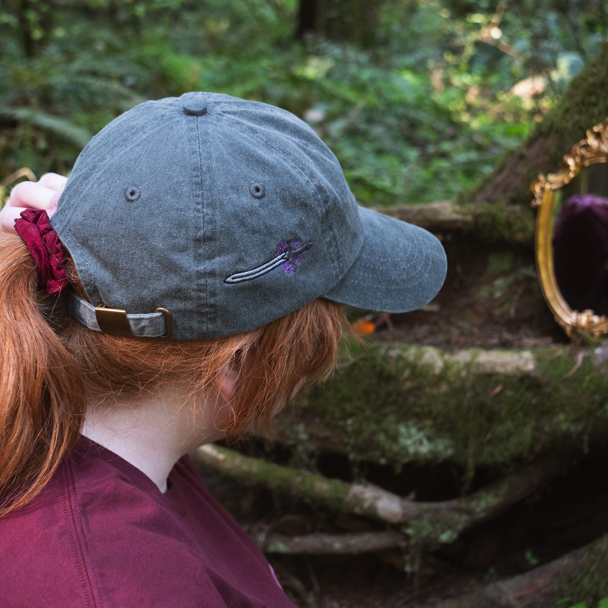 A model wearing the black Faithful Embroidered hat, looking at a gilded mirror in the forest.