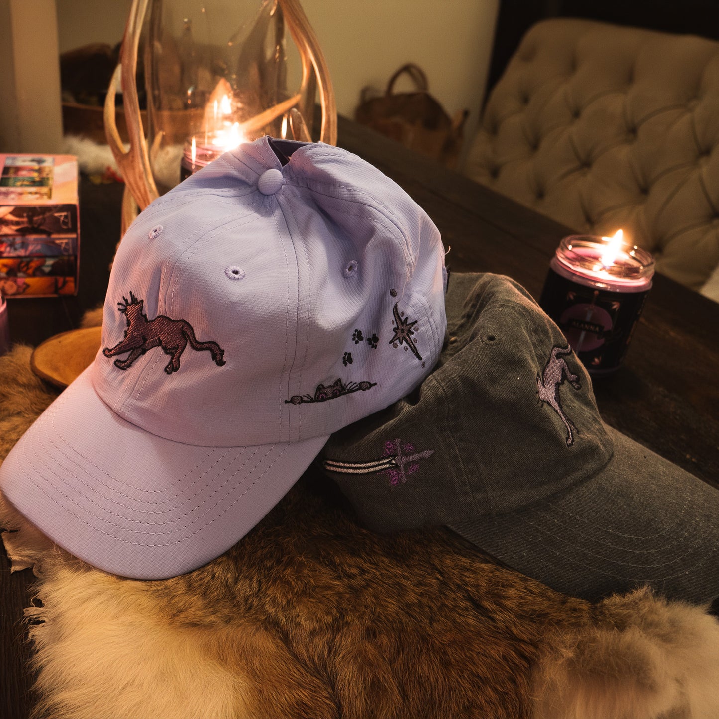 A photo of both the lavender faithful embroidered hat and the black faithful embroidered hat together on a wooden table, surrounded by lit candles, twinkling lights, the Alanna quartet, and a rabbit fur.