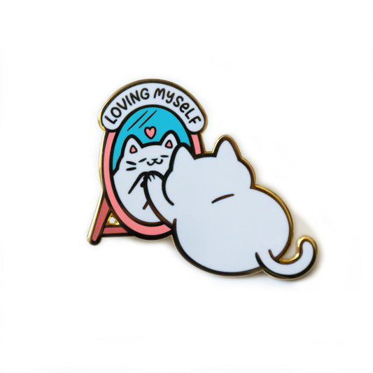 A product photo of an enamel pin depicting the back of a white cat facing a mirror with its paw resting on it. Reflected in the blue of the mirror is the cat’s chuffed face with a small pink heart above it. The mirror has a pink frame and the pin reads “Loving Myself” atop it with a white background.