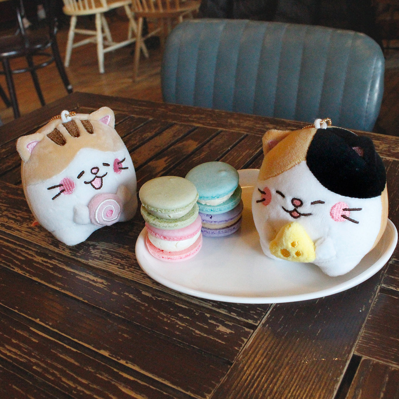 Two keyring plushes on a cafe table. Calico holds plush cheese while sitting on a plate with four multicolored macarons. Tan and white keyring cat sits beside the plate enjoying a plush pink cake roll.