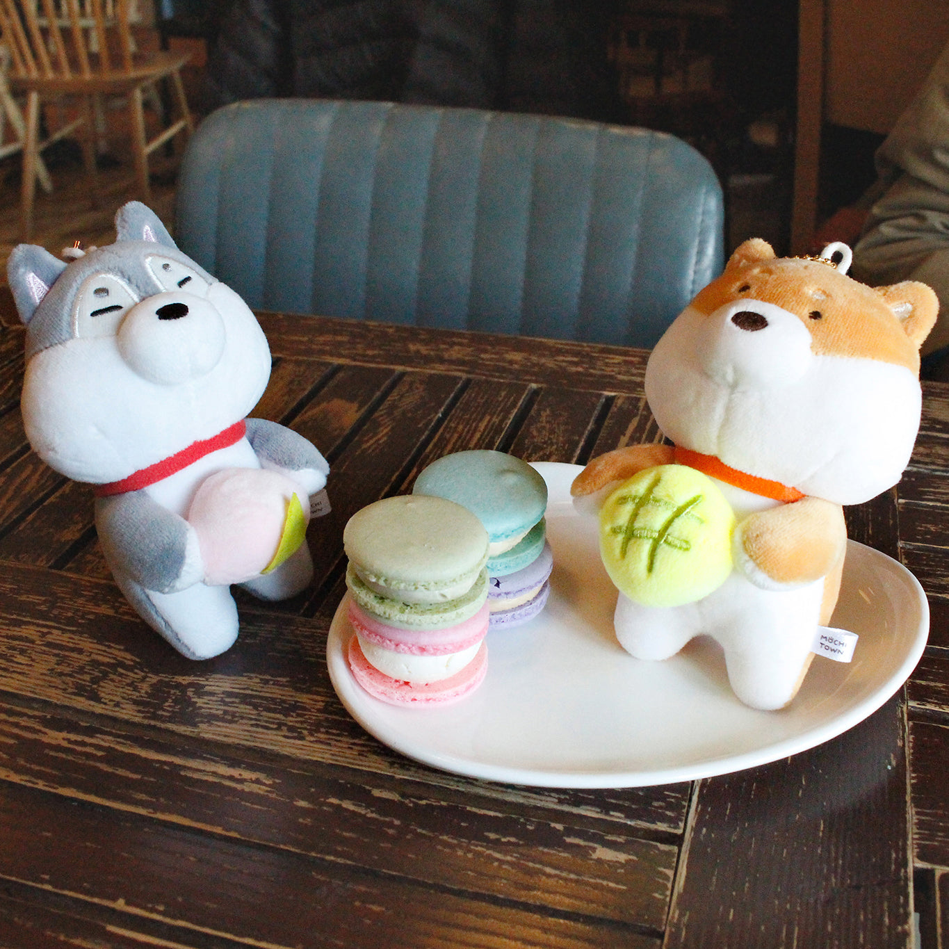 Shiba keyring and Husky keyring plushes on a cafe table. Tan shiba holds green plush roll while sitting on a plate with four multicolored macarons. Grey keyring shiba sits beside the plate enjoying a plush peach.