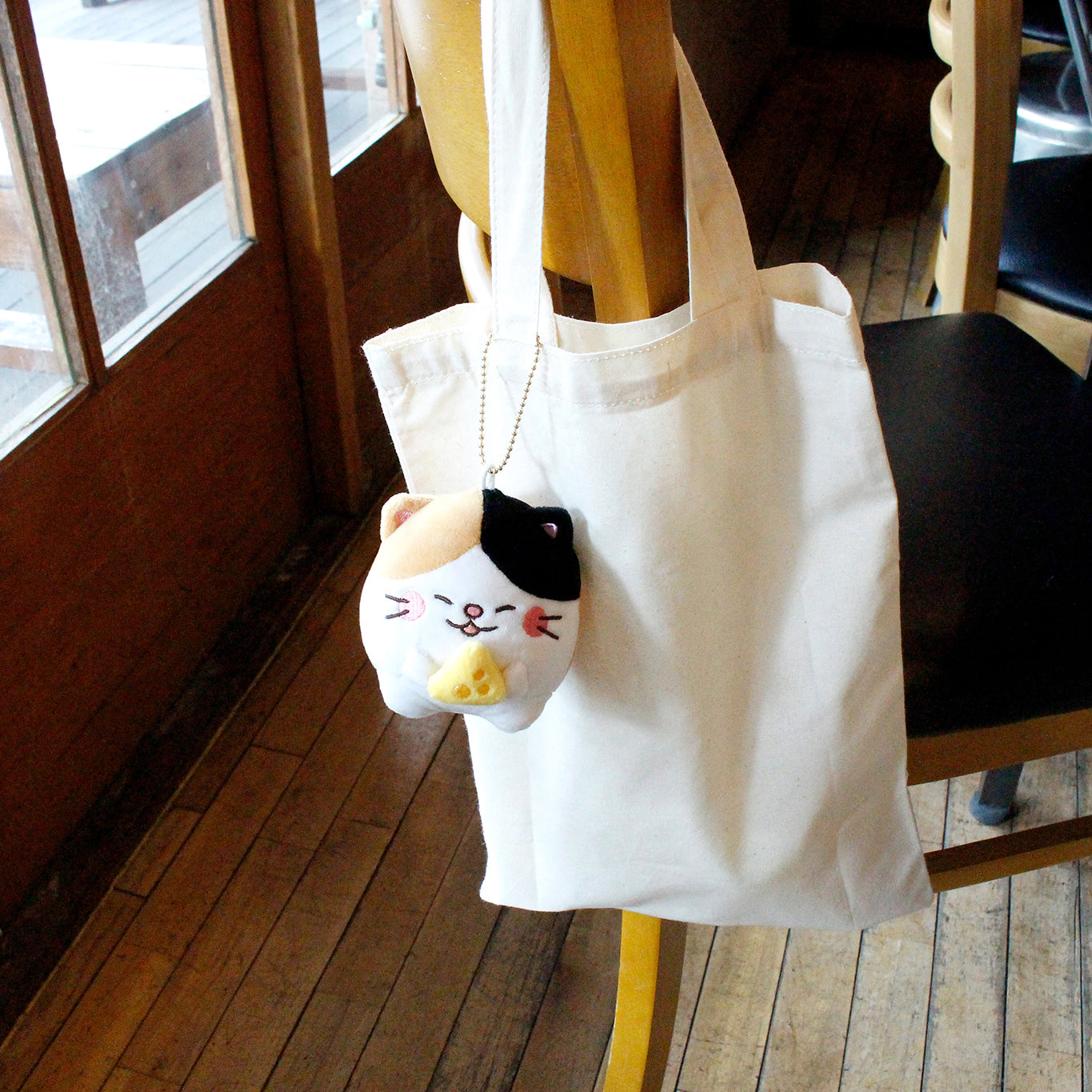 Calico cat with cheese attached to plain tote that is hanging off a cafe chair.