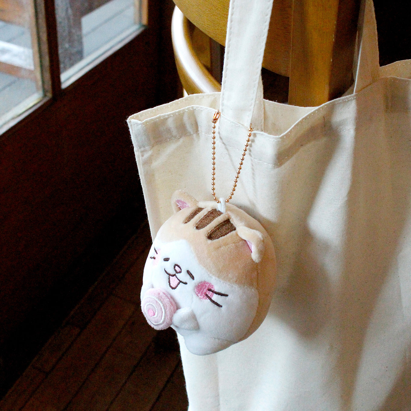 Beige cat holding pink cake roll keyring attached to plain tote that is hanging off a cafe chair.