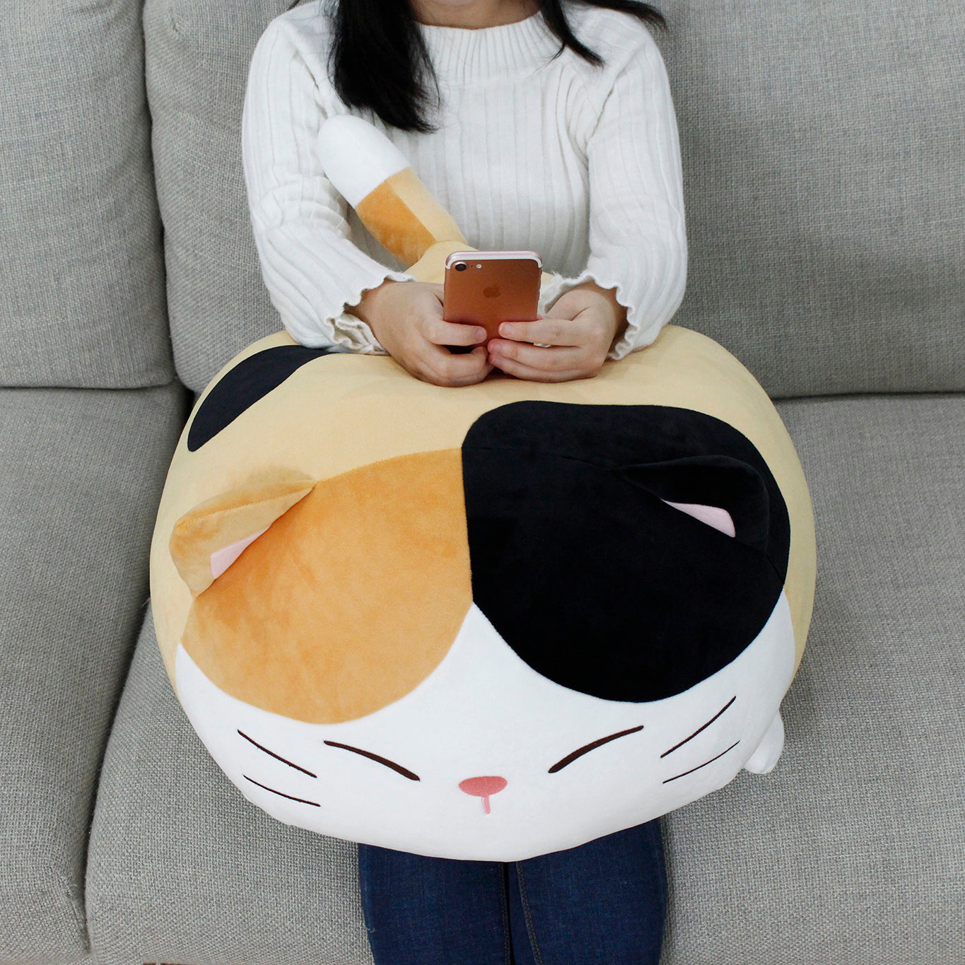 Front view of model sitting on grey couch using phone with Camang pillow on their lap, its tail sticking up.