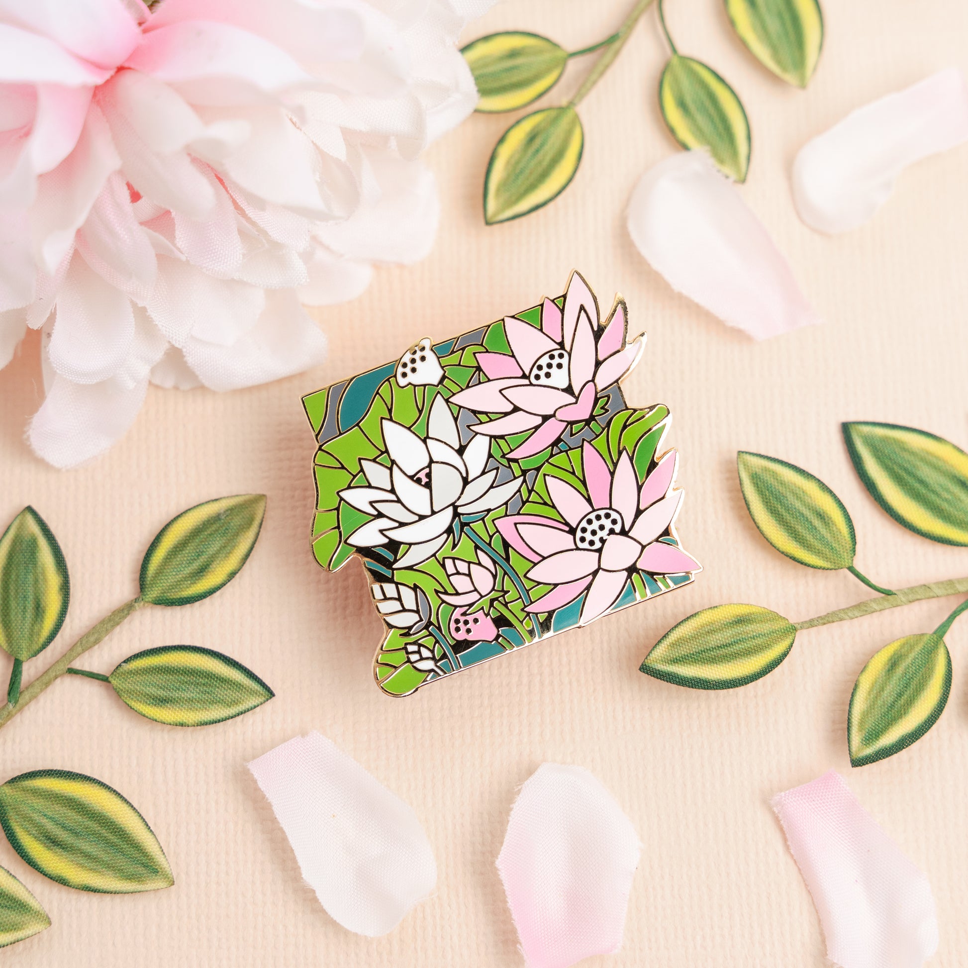 A lifestyle photo of the Demigirl Lotus Pin laying on a peach table with pink flowers and flower petals that match the pin's pastel pink and white colors.