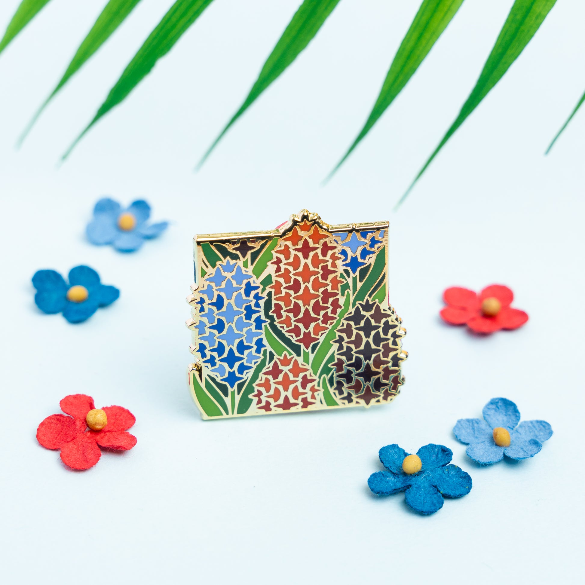 A lifestyle photo of the Polyamory Hyacinths Pin standing surrounded by red and blue paper flowers. The floral elements compliment the pin's orange, brown, green, and blue colors.