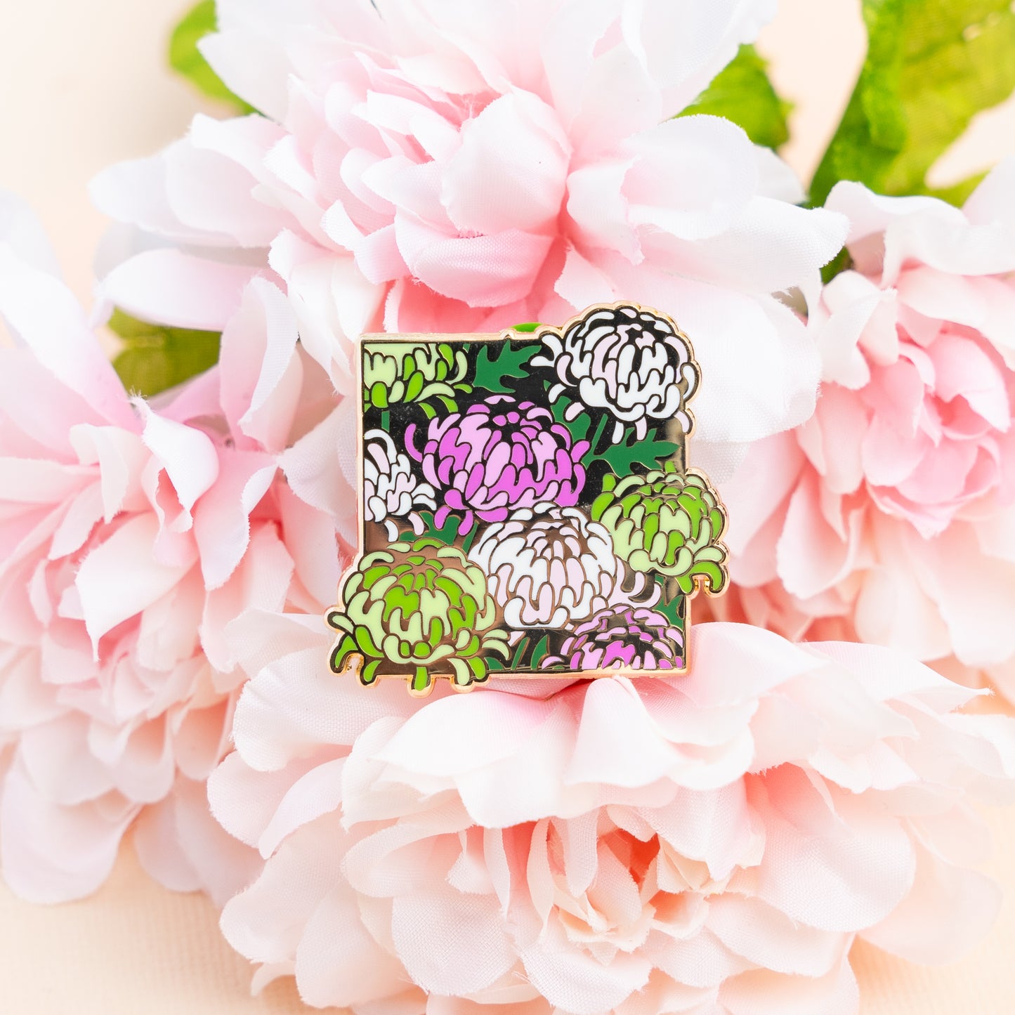 A lifestyle photo of the Genderqueer Chrysanthemums pin showing off its pinks and greens from atop a bundle of peony flowers.