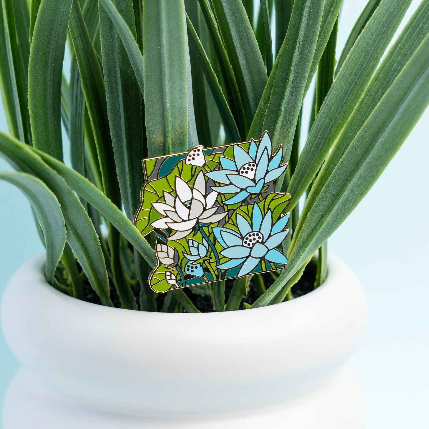 The blue, white, and green Demiboy Lotus pin is aesthetically pinned to a fake plant in a white planter.