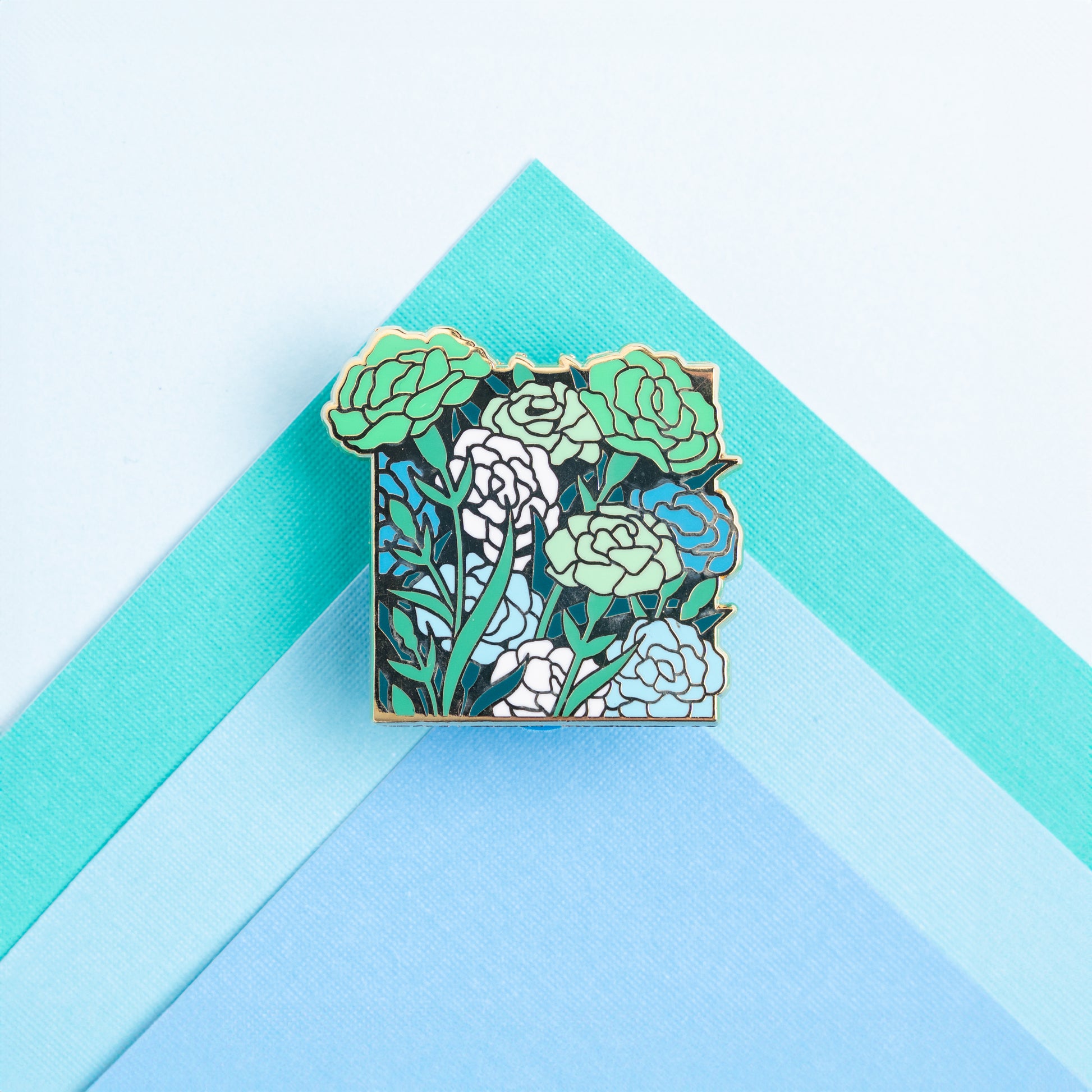 A lifestyle photo of the Achillean Carnations pin pinned to papers of matching green and blue colors.