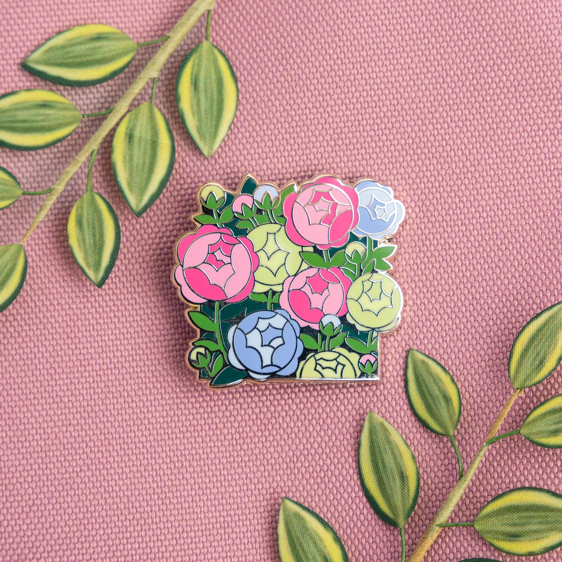 A lifestyle photo of the Poly Peonies Pin on a pink fabric surrounded by green leaves that compliment the pin's pink, green, and blue colors.