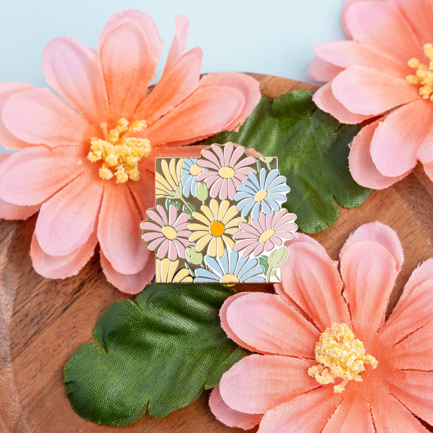 A lifestyle photo of the Pan Daisies pin amidst fake peach-colored flowers that compliment the pin's pastel pink, yellow, blue, and green hues.