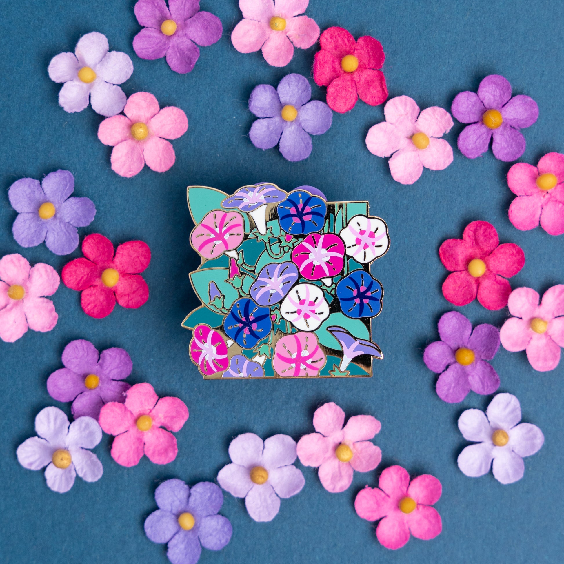 The colorful Genderfluid Morning Glory pin on a teal table amongst small felt flowers that match the pin's pink, purple, blue, and teal green hues.