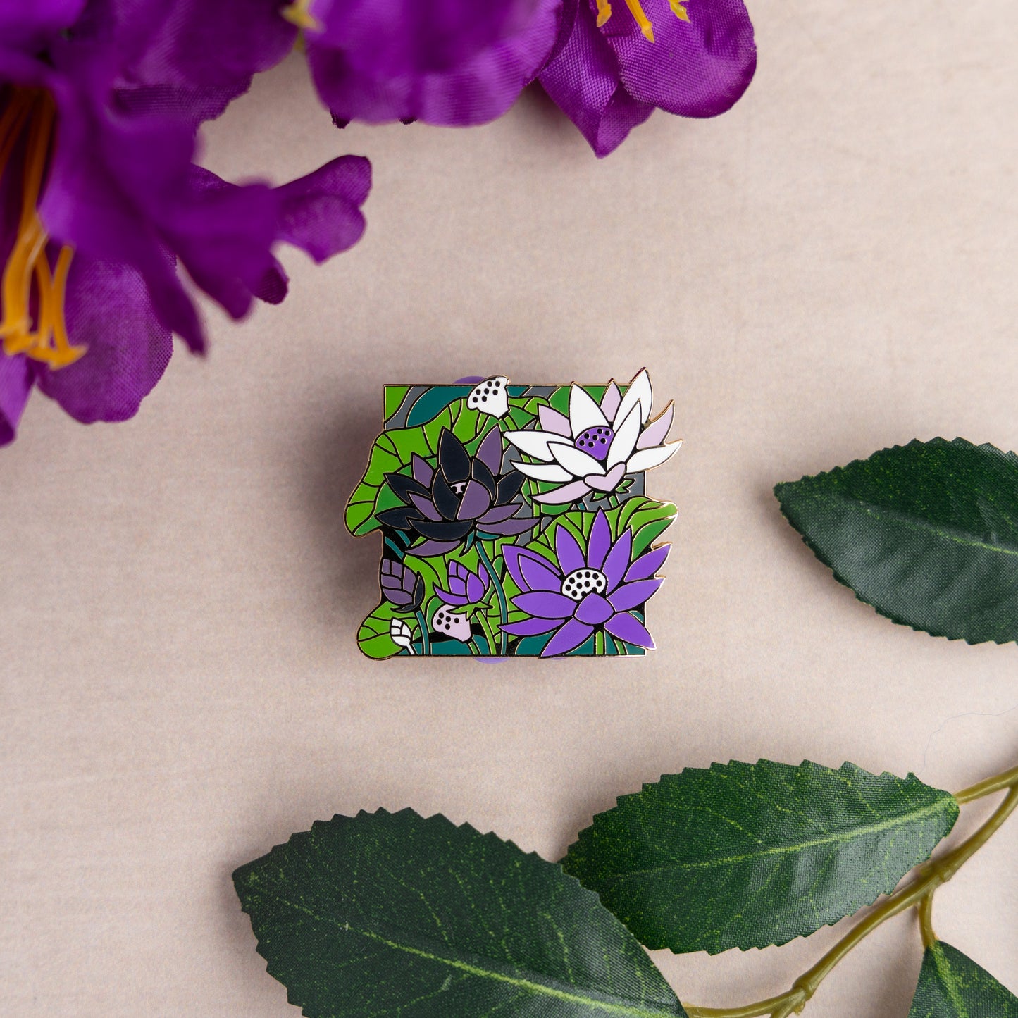 A lifestyle photo of the Demi Lotus Pin on a table amidst matching purple flowers and green leaves.