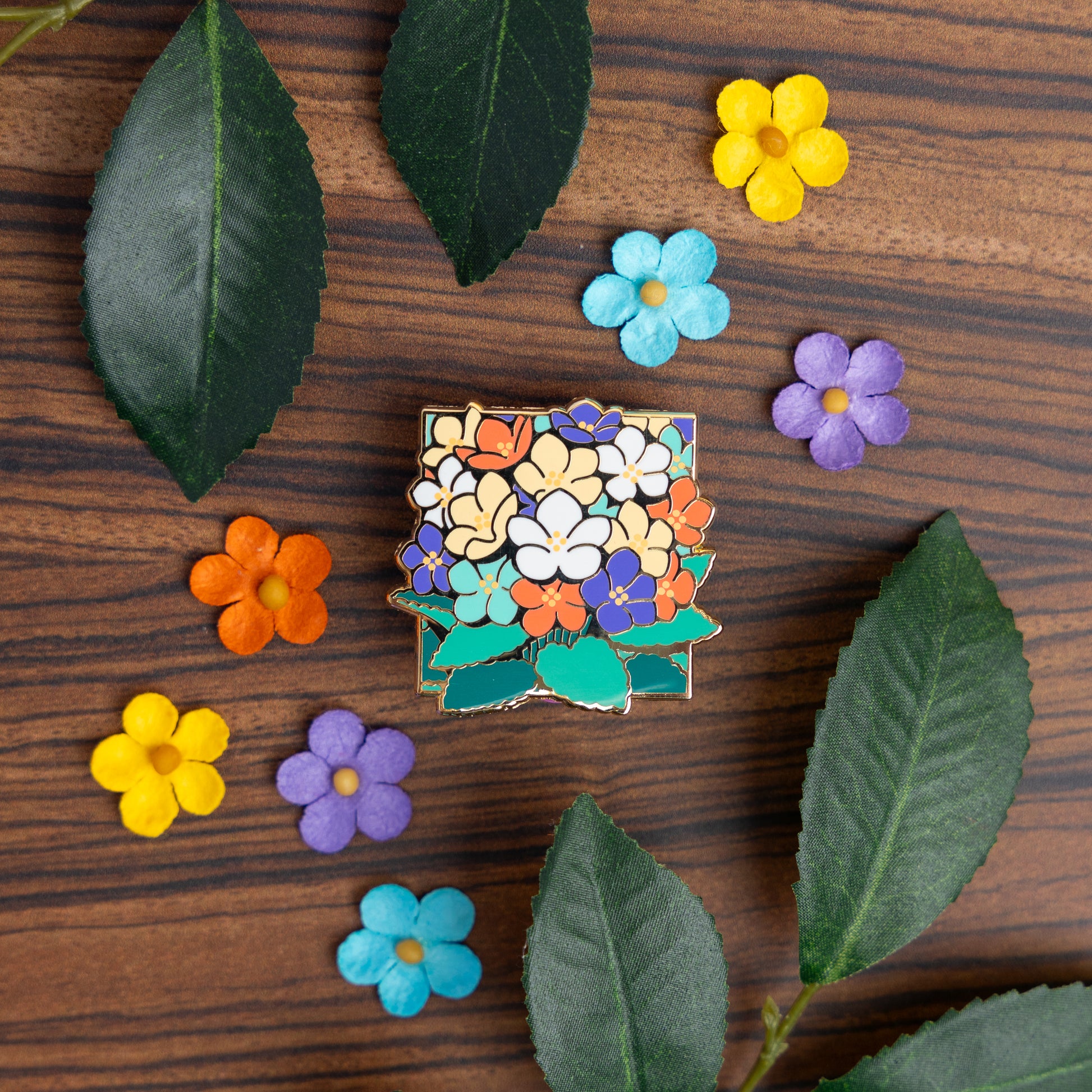 A lifestyle photo of the Sapphic Violets Pin laying on a table surrounded by paper flowers and leaves that match the pin's buttery yellow, white, violet, orange, and teal green color scheme.