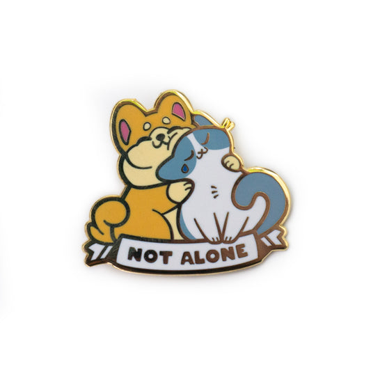 A product photo of an enamel pin depicting a yellow shiba inu dog hugging a grey and white cat. The shiba inu’s face is squished against the cat’s face. The cat is serenely crying and looks comforted. They are sitting on a horizontal white ribbon that reads “Not Alone.”