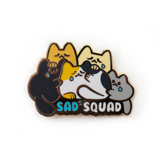 A product photo of an enamel pin that shows five blobby cats gathered together. From left to right: black, orange, cream, calico, and grey cats. Four of them have a small blue tear coming from one eye. Two of their tails hang down to frame the text on the pin: “Sad Squad.”
