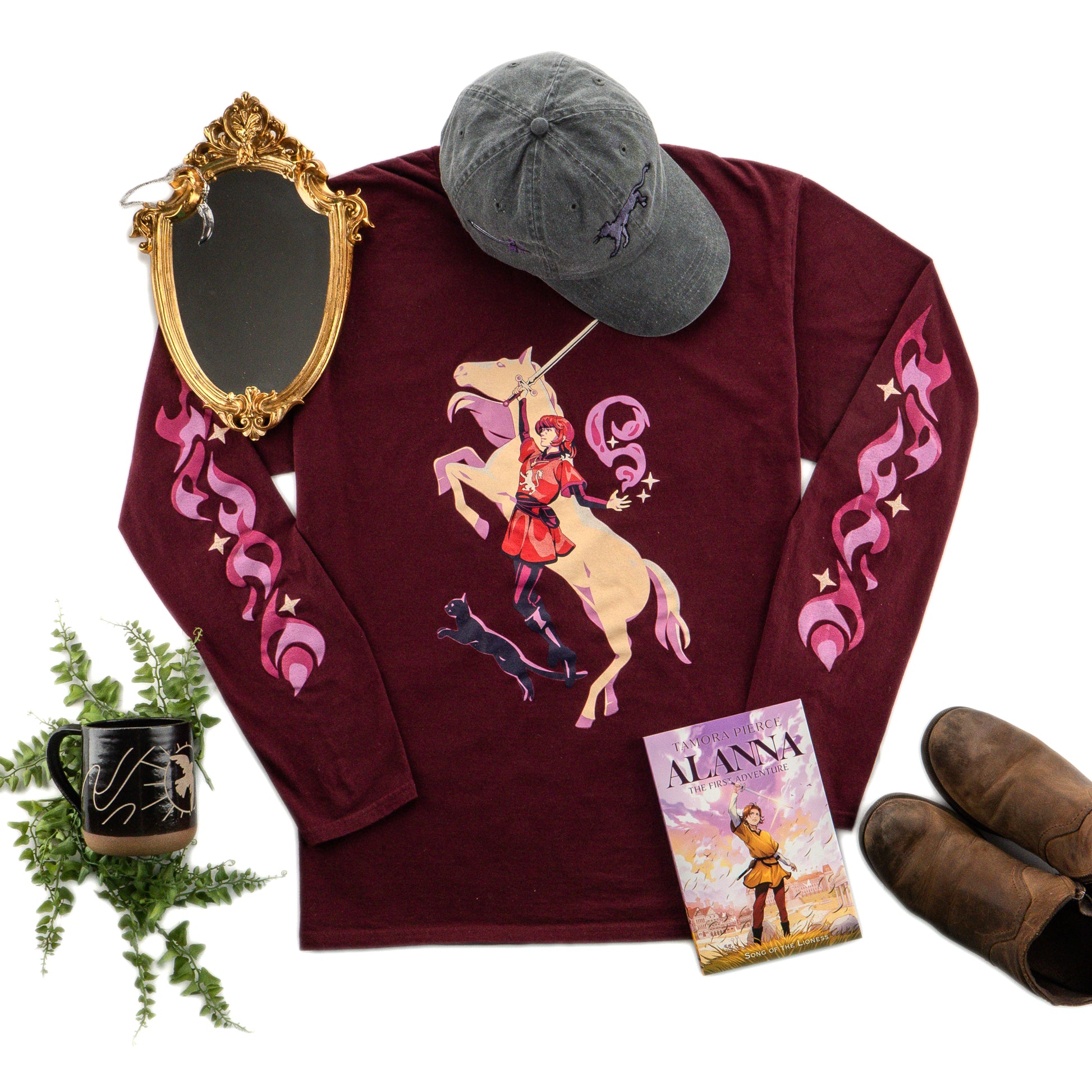 the maroon t-shirt is laid flat, surrounded by brown boots, an Alanna book, a black ballcap, a mirror, and foliage.