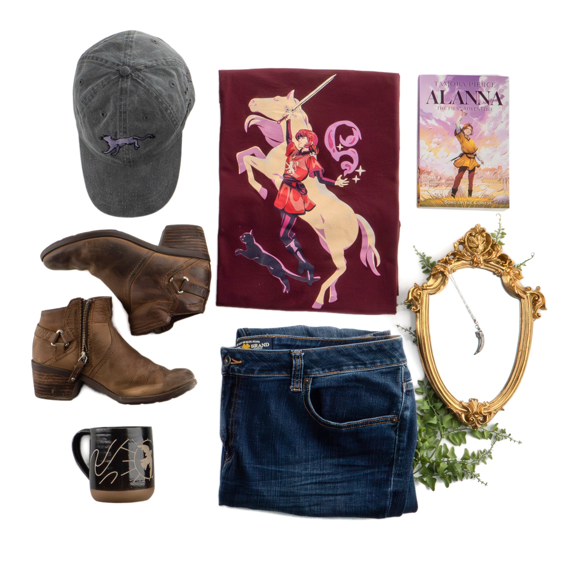 the long-sleeve t-shirt is folded into a neat rectangle, surrounded by a black ballcap, brown boots, a folded pair of denim jeans, the Dancing Dove mug, an Alanna book, a mirror with the badger claw pendant laid on top, and some foliage.