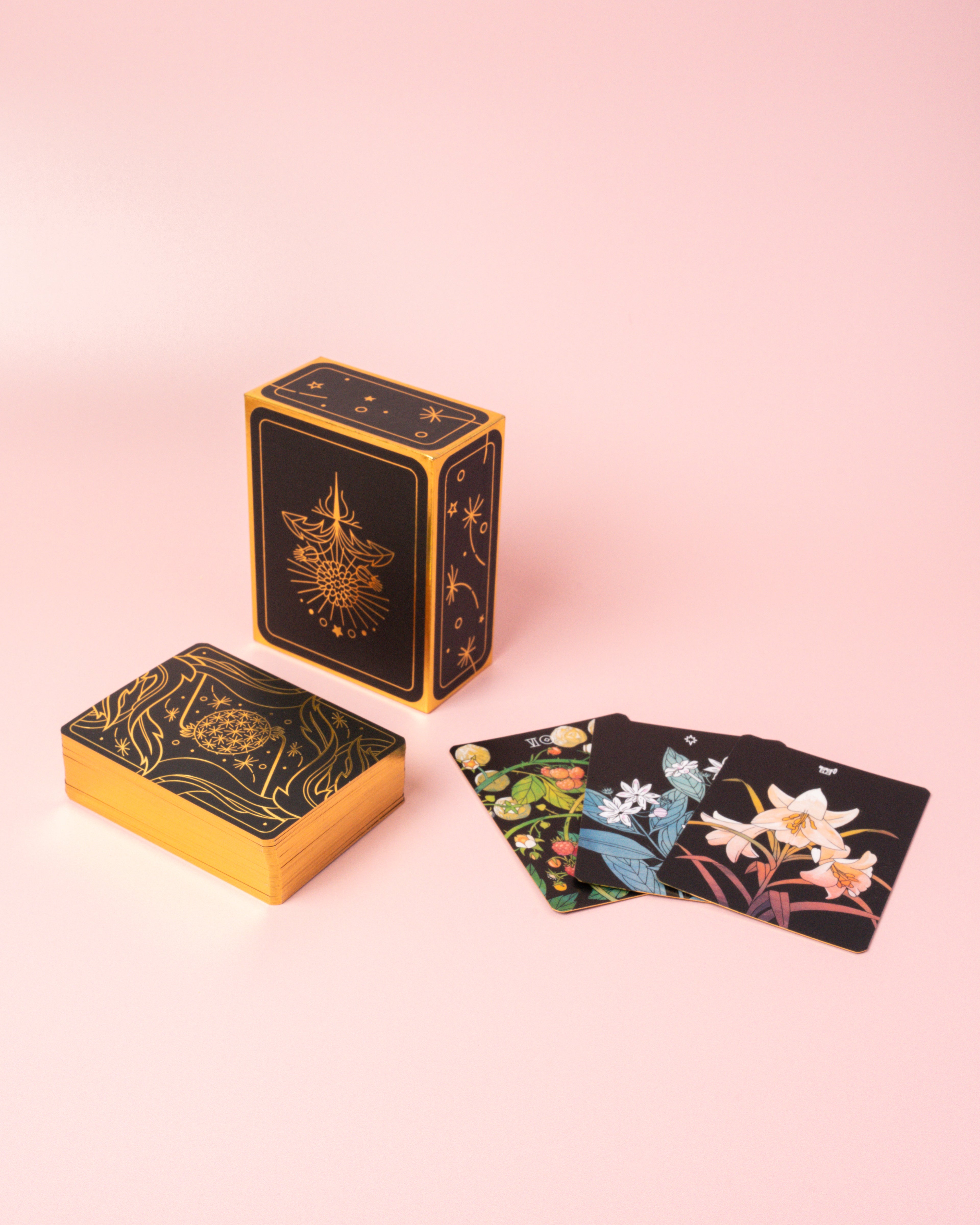 An intricately designed black and gold Botanica tarot deck box with floral and astral motifs, a face-down stack of decorative and golden-edged tarot cards, and a three-card spread of face-up cards with flower illustrations on a blush pink background.  