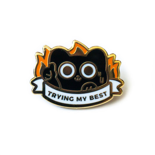 A product photo of an enamel pin depicting a black cat’s head and front paws peeking over a white ribbon that says “Trying My Best.” The cat is sweating, giving a thumbs up, and flames are creeping up behind it.