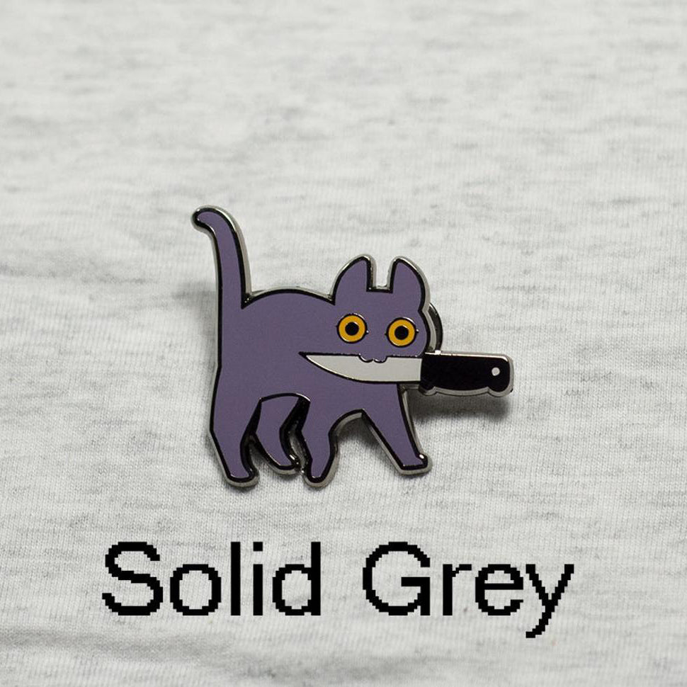 Silver enamel pin of grey cat with orange eyes holding knife in mouth with heather grey cloth background. Caption text reads: "Solid Grey."