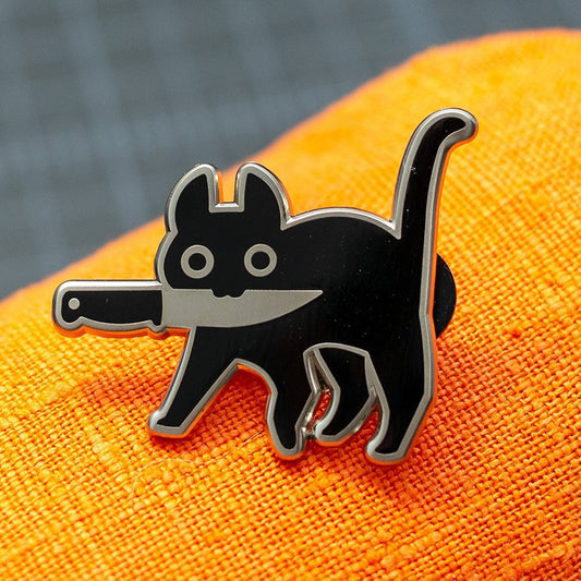 Silver enamel pin of black cat holding knife in mouth with orange cloth background.