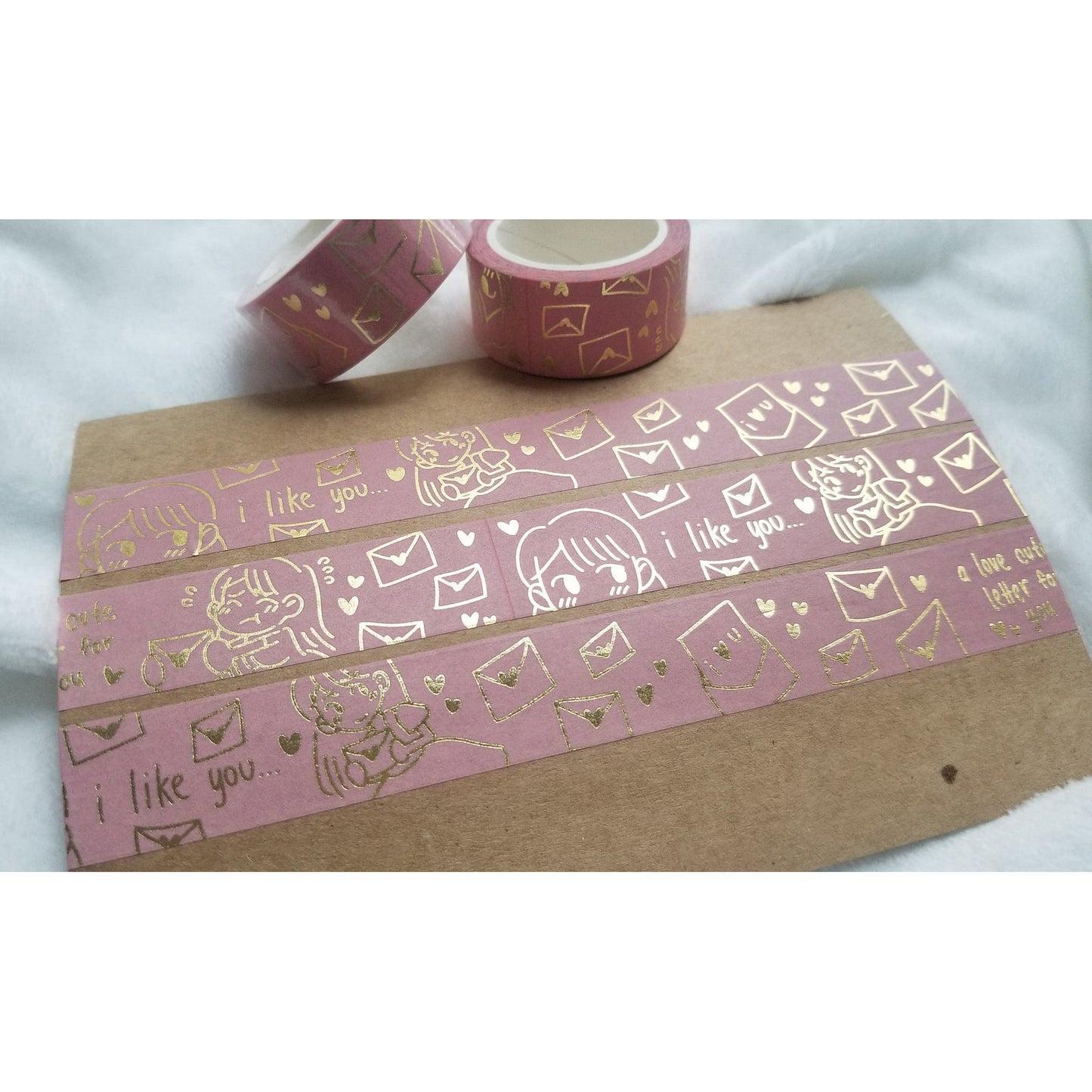 Rose pink with gold foil washi tape design of hearts and envelopes with a blushing person. Three strips of the tape on thin board on a white blanket with two full rolls of tape.