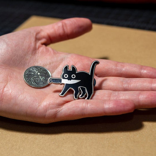 Silver enamel pin of black cat holding knife in mouth with coin for scale in model's hand.