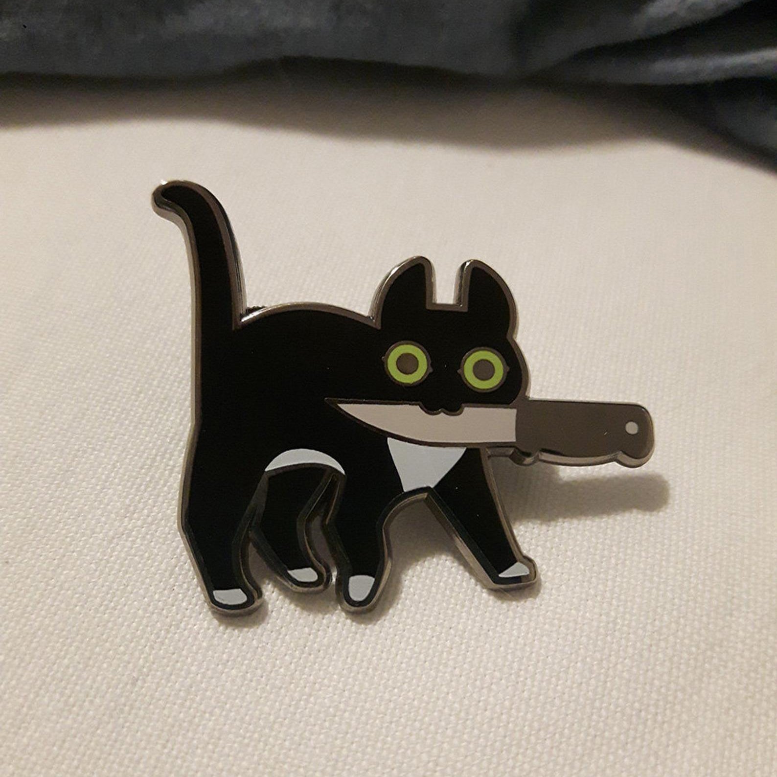 Silver enamel pin of tuxedo cat with green eyes holding knife in mouth with white cloth background.