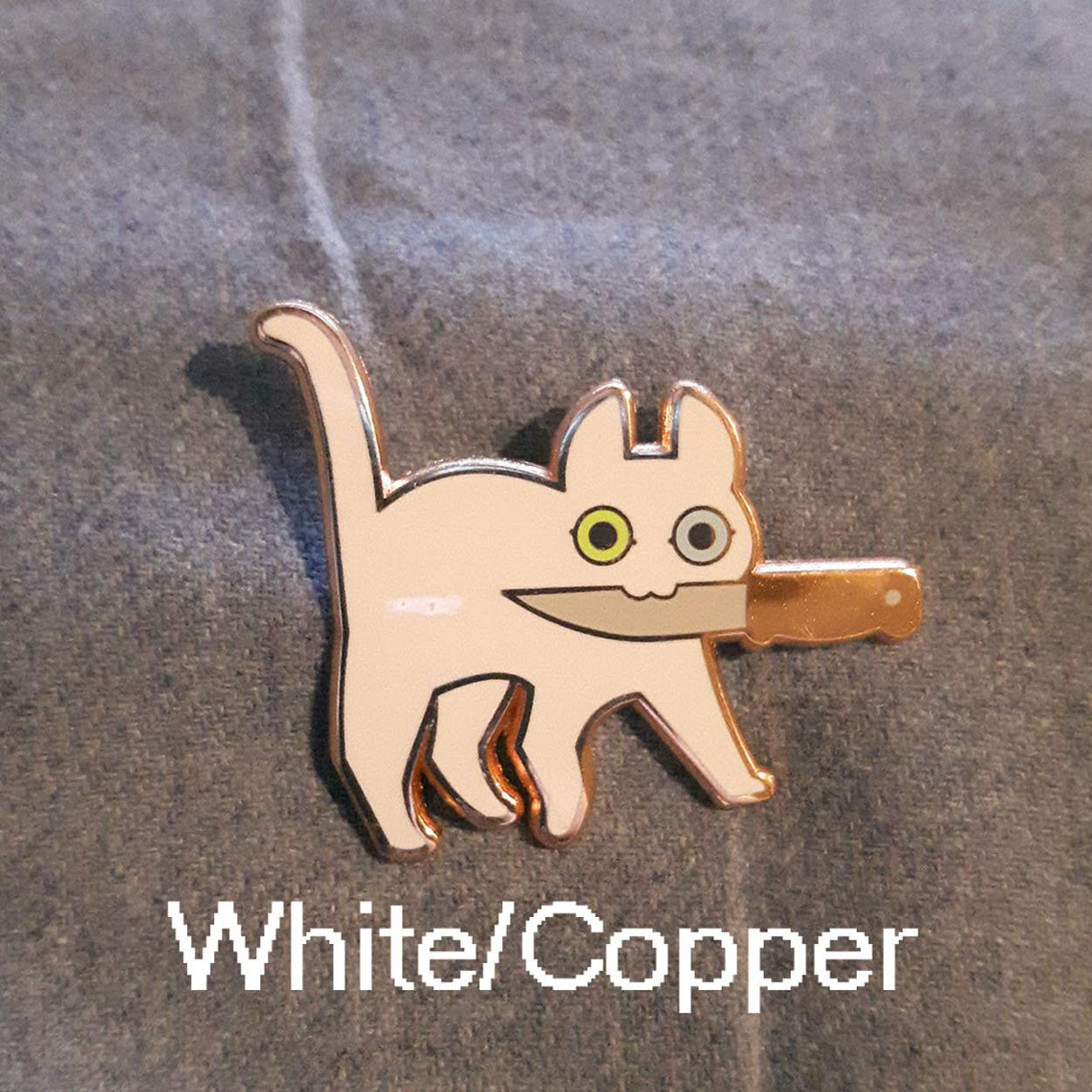 Alternate white and copper version of black cat holding knife in mouth enamel pin on grey cloth background.