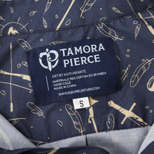 Load image into Gallery viewer, Detail of dark blue sewn-in Tamora Pierce logo tag, with art credit to Kaiti Infante, and material contents and care.