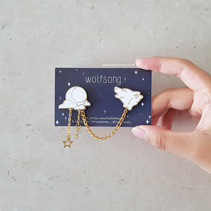 A hand holds the white variation of Wolf & Moon collar pins. White wolf howling enamel pin is connected to white moon and clouds by golden delicate chain with tiny star charm.