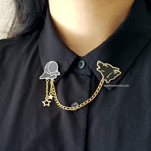 The white variation of Wolf & Moon collar pins on model wearing black button up. Black wolf howling enamel pin is connected to black moon and clouds by golden delicate chain with tiny star charms.