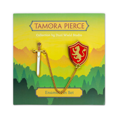Front view of the Alanna Sword & Shield Pins on forest backing card. The Gold enamel pins have red and white details and are connected by a delicate gold chain.