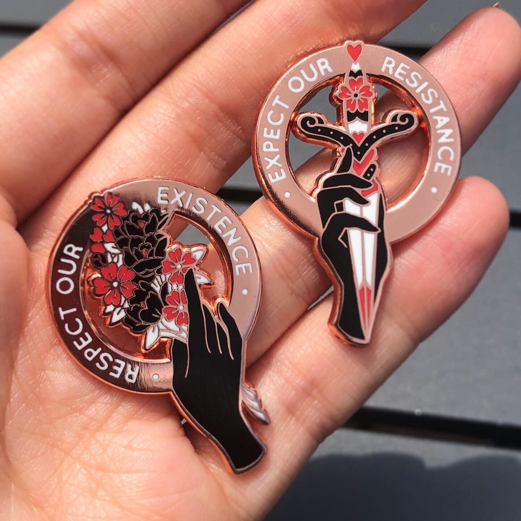 Two rose gold enamel pins held in a hand. Left enamel pin reads 