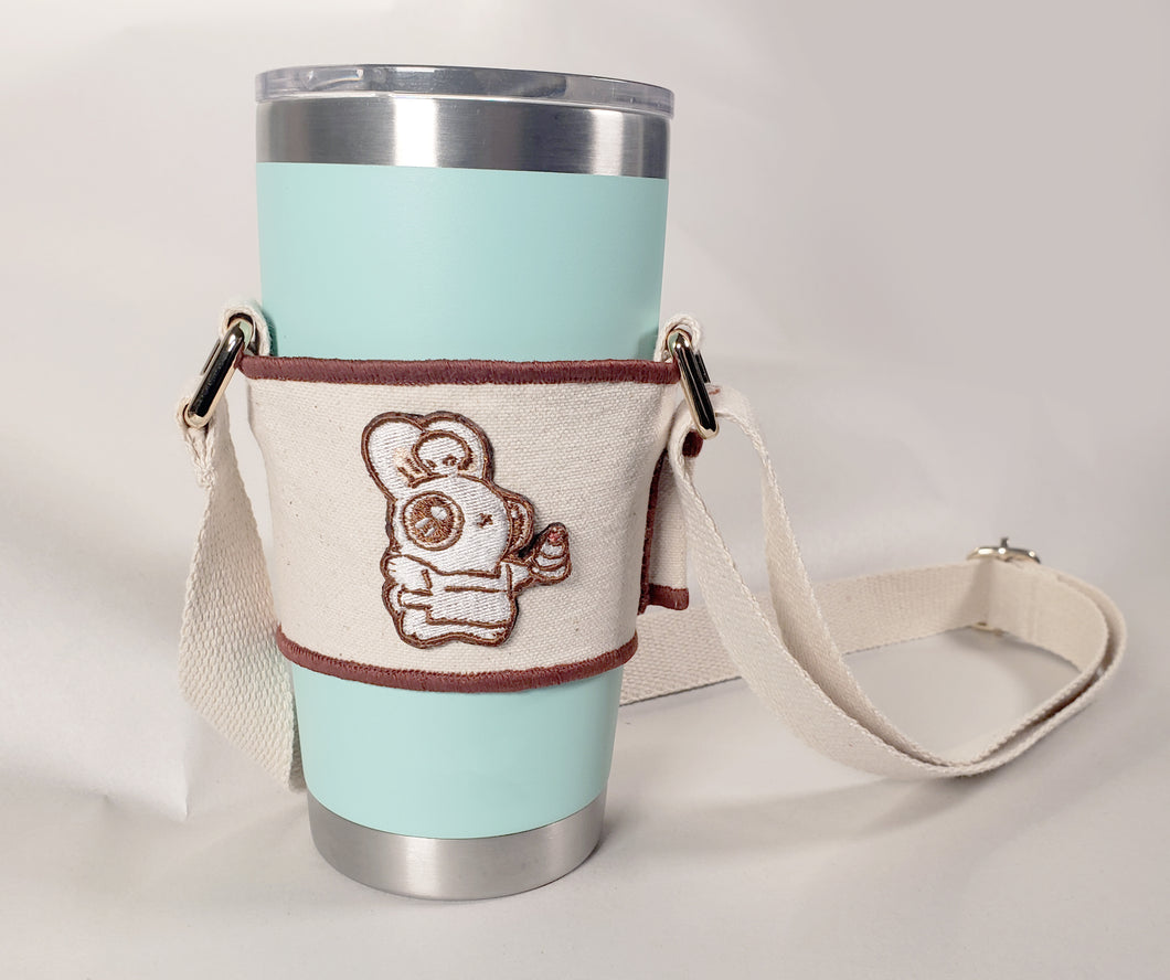 Linen color cloth cup holder with Baker Bunneh as an embroidered patch holding a mint green coffee tumbler.