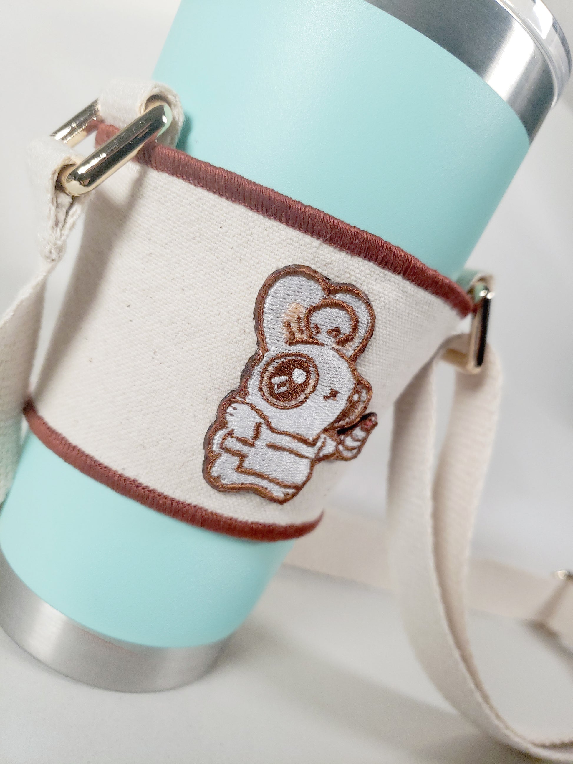 Linen color cloth cup holder with Baker Bunneh as an embroidered patch holding a mint green coffee tumbler.