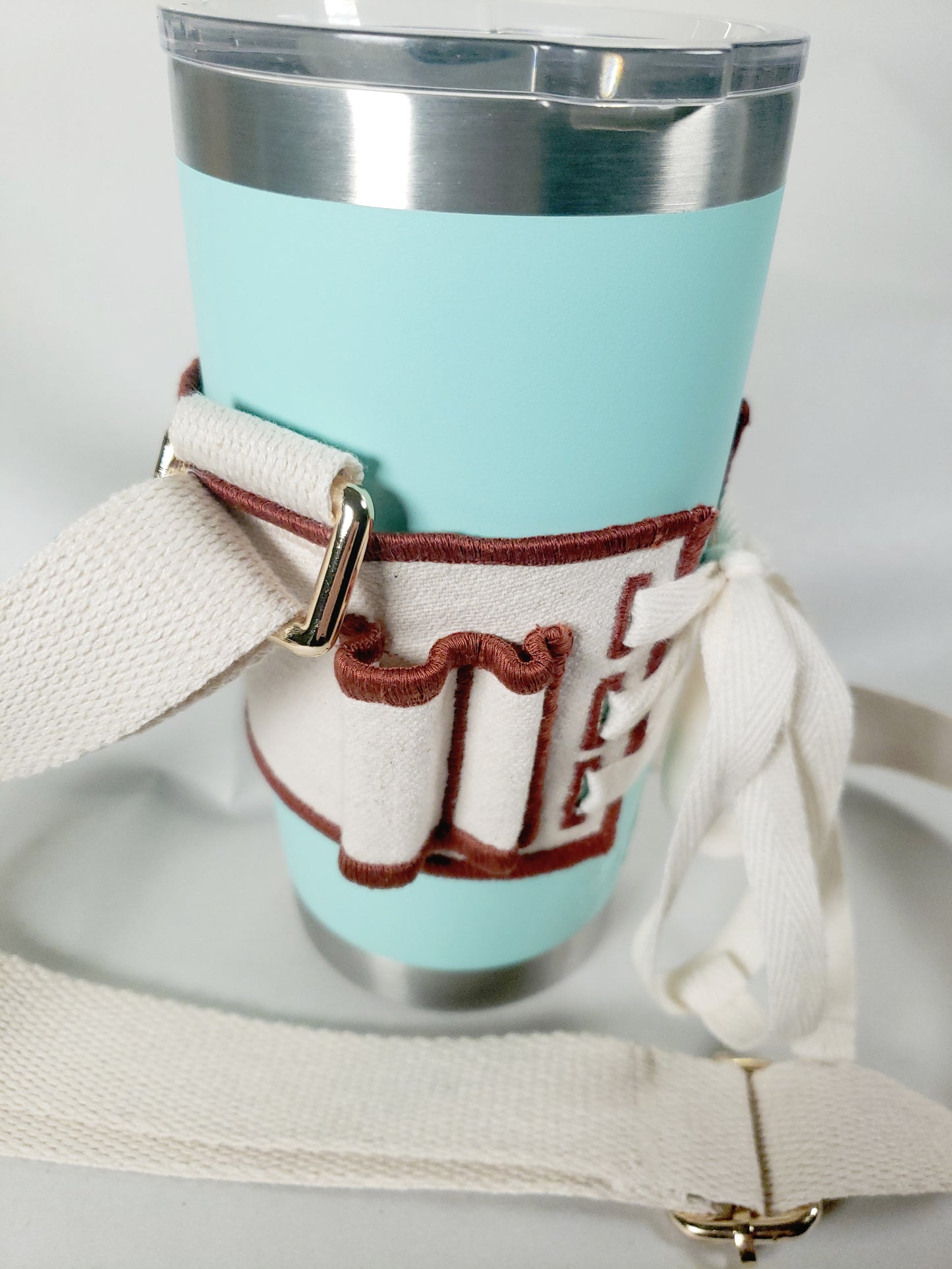 Closeup of Linen color cloth cup holder with Baker Bunneh as an embroidered patch holding a mint green coffee tumbler.