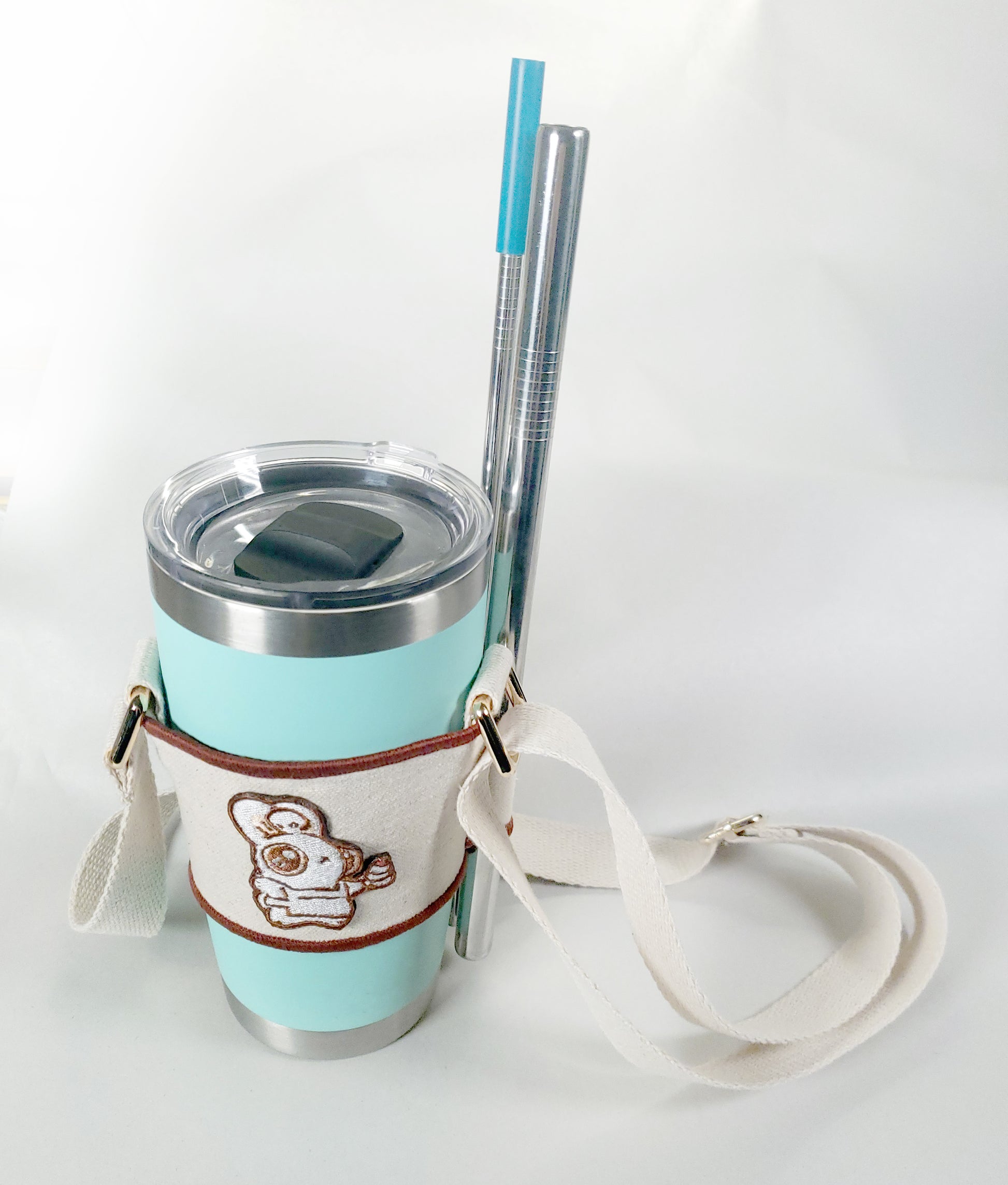 Linen color cloth cup holder with Baker Bunneh as an embroidered patch holding a mint green coffee tumbler and metal straw..
