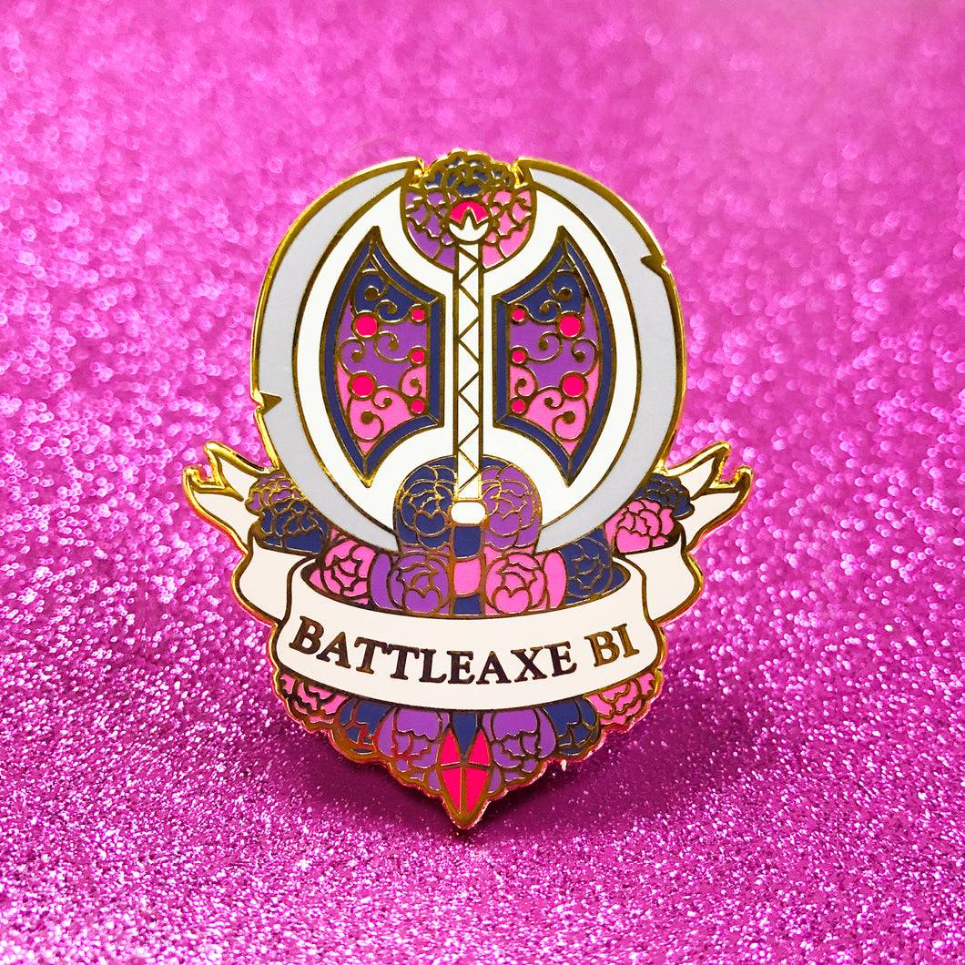 The goldcast pin features a dual bladed axe and roses in the colors of the bisexual flag. A ribbon trails across the front and reads 