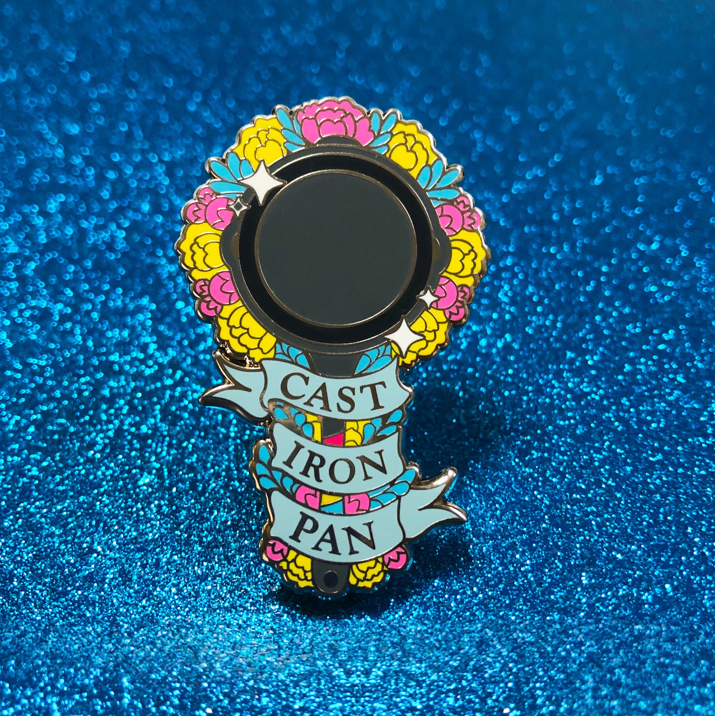 The goldcast pin features a cast iron cooking pan and flowers in the colors of the pansexual flag. A ribbon trails across the front and reads "Cast Iron Pan"