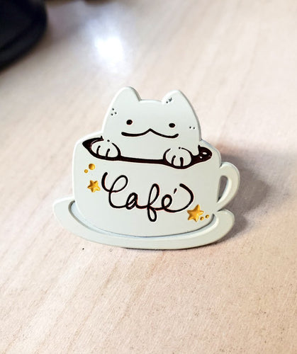 Enamel pin of stylized, smiling white cat peeking out with paws from a full coffee cup and saucer that says 