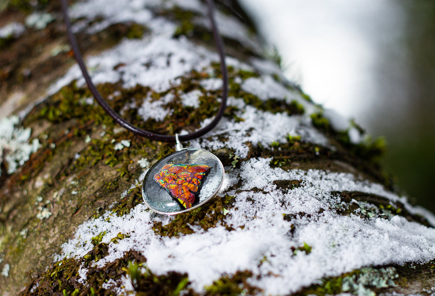 A molten lava-like looking stone with azure details floating in a gold-rimmed circular pendant. Pendant is on brown necklace cord resting on a snow-dusted, mossy tree log.