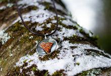 Load image into Gallery viewer, A molten lava-like looking stone with azure details floating in a gold-rimmed circular pendant. Pendant is on brown necklace cord resting on a snow-dusted, mossy tree log.