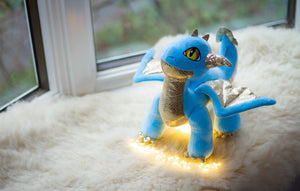 Skysong plush stands in window alcove on plush white blanket with fairy lights at her feet.